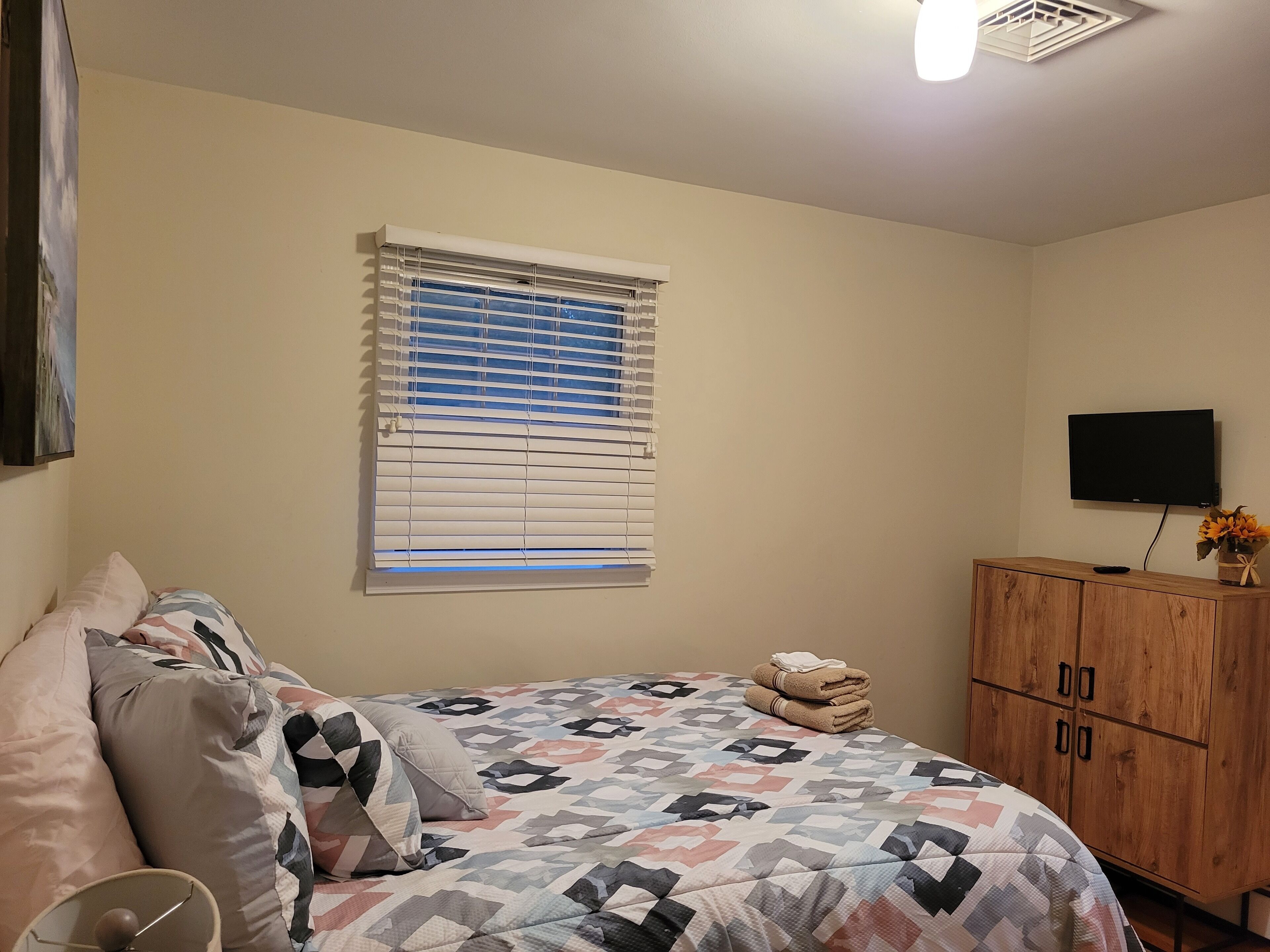 Une chambre à coucher avec un lit, une fenêtre avec des stores, un téléviseur fixé au mur et une armoire en bois.