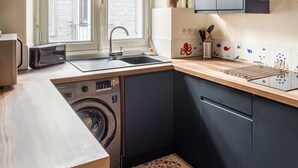 A compact kitchen with a washing machine, a sink, and a window with a view of a building. - Le Traou Mad - Au cœur de l'Intra-muros (Saint-Malo)