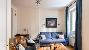 A living room with a blue sofa, a wooden coffee table, and a wicker chair. There is a painting on the wall and a chandelier. - Le Traou Mad - Au cœur de l'Intra-muros (Saint-Malo)