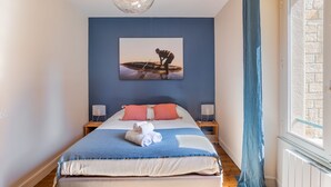 A bedroom with a bed, a painting, bedside lamps, and a window with curtains. - Le Traou Mad - Au cœur de l'Intra-muros (Saint-Malo)