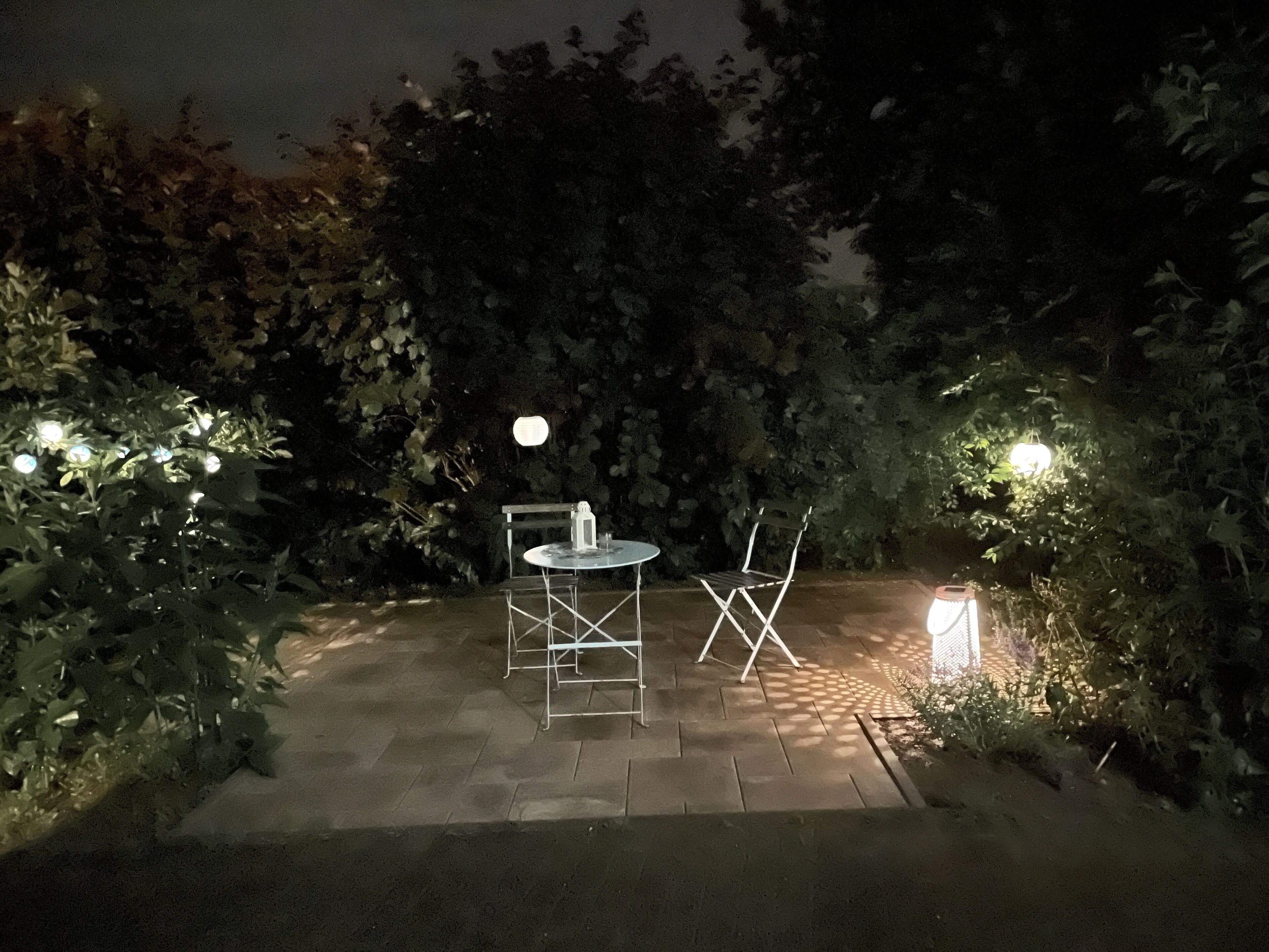 An outdoor patio area with a round table and two chairs, surrounded by greenery and illuminated by small lights.
