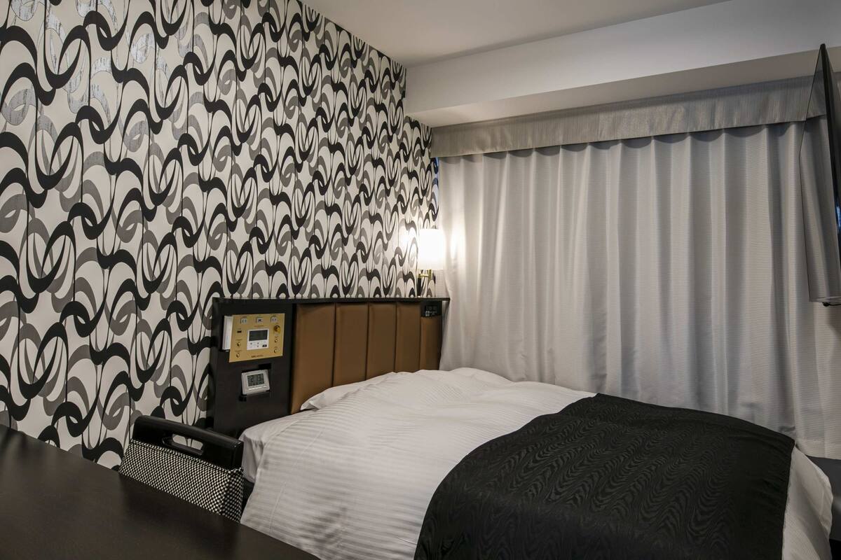 Ein Hotelzimmer mit ornamentalem Wandpapier, einem Bett mit weiß-schwarzem Überwurf, einem Holz-Kopfteil, einem Nachttisch mit Lampe und einem Fenster mit Vorhängen.