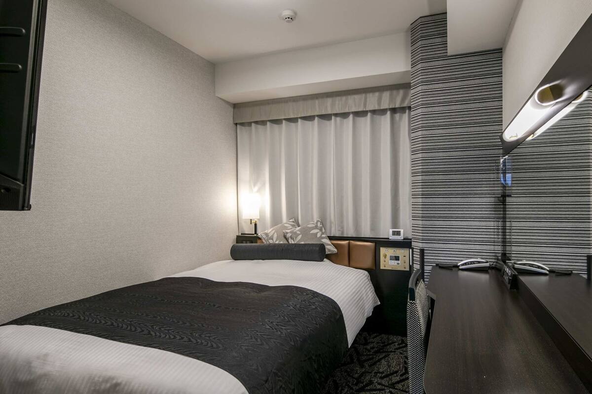 Ein Hotelzimmer mit zwei Betten, einem Schreibtisch, einer Lampe, einem Fenster mit Vorhängen und einer Deckenleuchte.