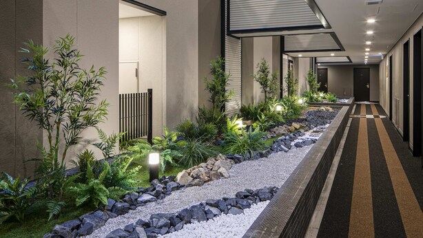 Un couloir d’hôtel moderne, avec un long passage étroit bordé de plantes et de rochers, qui mène à une porte au bout.