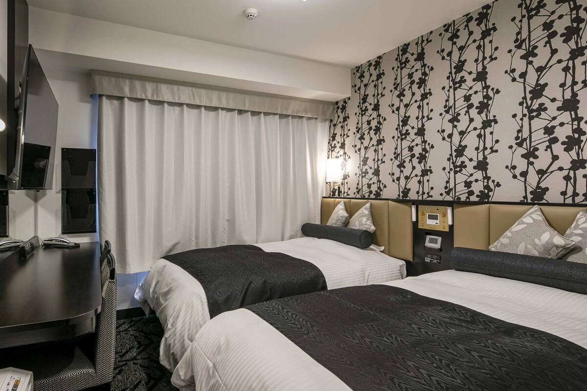 Ein Hotelzimmer mit zwei Betten, einem Schreibtisch mit Telefon, einem Fernseher und einer an der Wand montierten Steuerung.