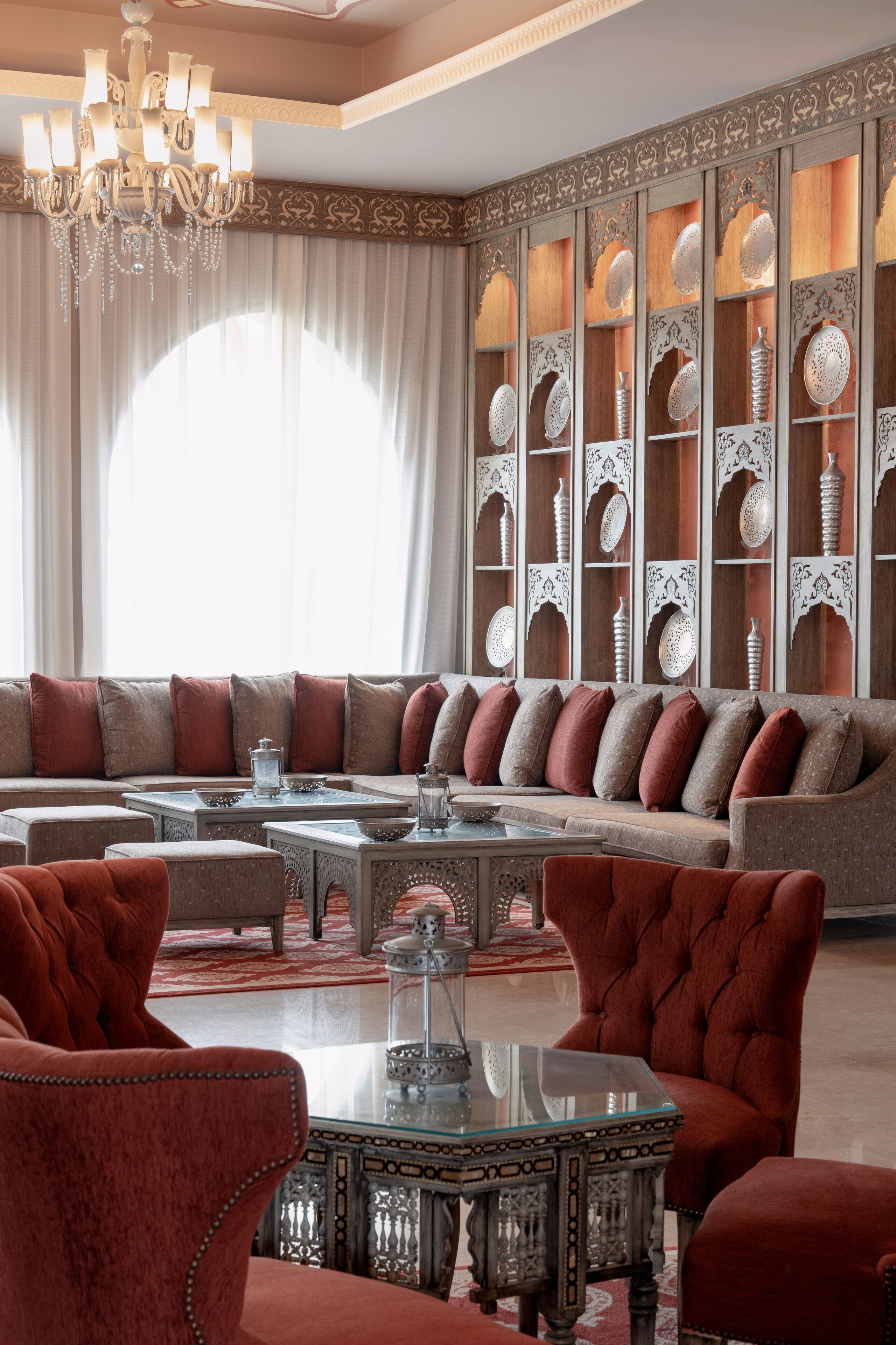 Un salon luxueux avec un grand canapé, des fauteuils rouges, une table basse en verre et un mur décoratif agrémenté d’étagères.