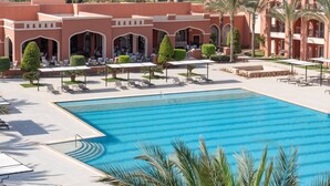 A large swimming pool with lounge chairs and umbrellas, surrounded by a landscaped area with palm trees and a building with arches. - JAZ Tamerina, Almaza Bay (Marsa Matrouh)