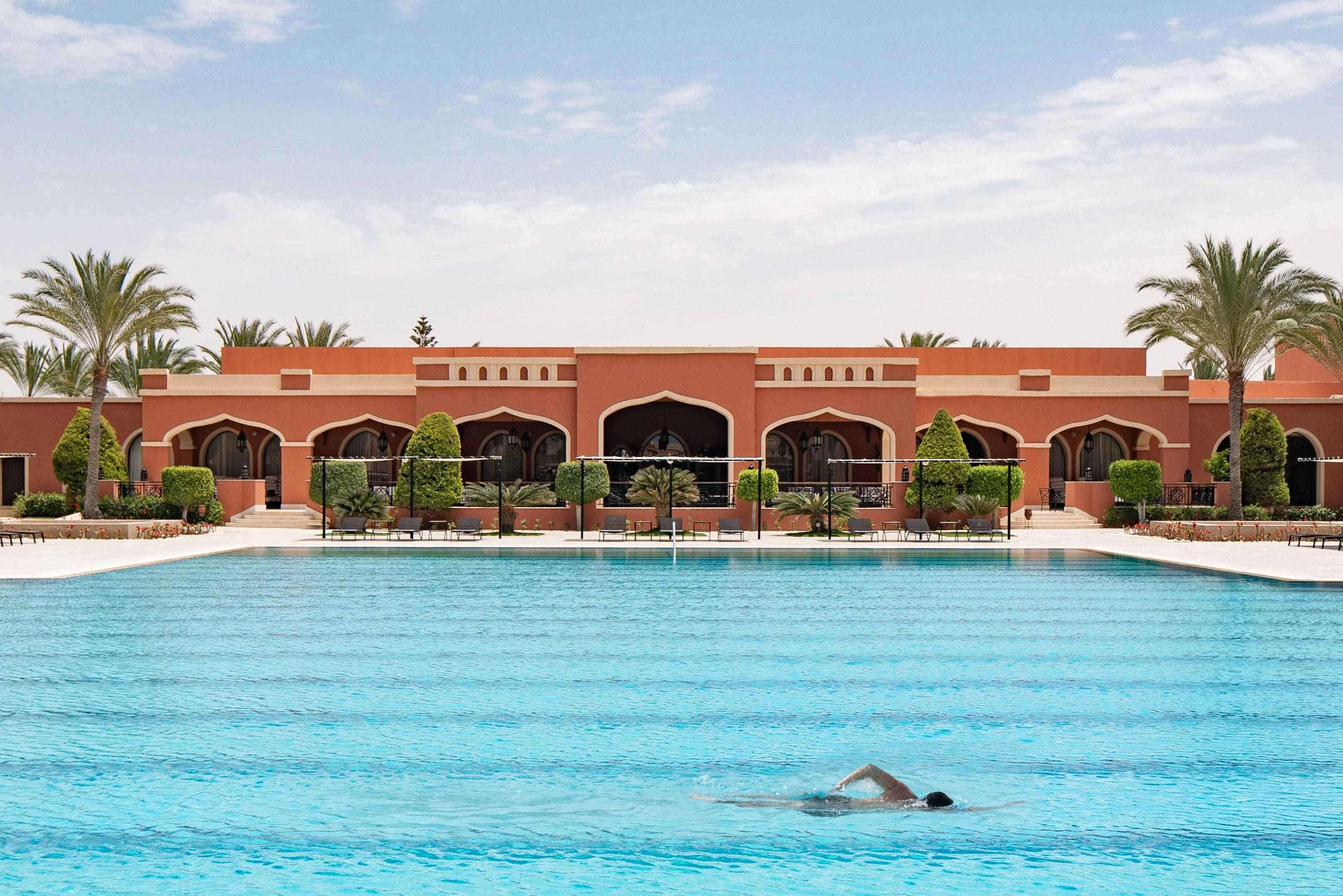 Piscina com uma pessoa nadando, cercada por um edifício com arcos e palmeiras.