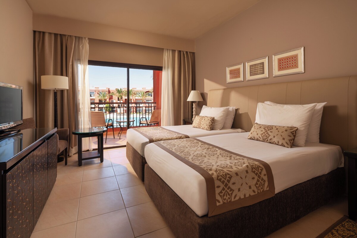A hotel room with two beds, a TV, a desk, and a balcony with a pool view.