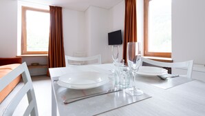 Hotelkamer met een eettafel gedekt voor twee personen, twee witte stoelen, een flatscreen televisie aan de muur en grote ramen met gordijnen.
