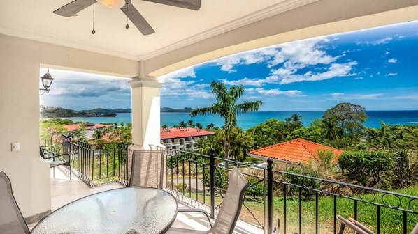 Un balcon avec une table et des chaises en verre, donnant sur un paysage côtier sous un ciel dégagé.