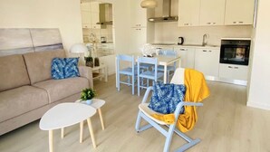 A modern living room with a sofa, coffee table, and rocking chair. A kitchen with white cabinets and appliances is visible in the background. - Apartment on the beach 300 meters from the sea, wifi, pool and tennis, ideal for families (L'Escala)
