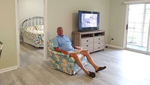 A man sitting on a floral-patterned chair in a room with a television on a stand, a bed with a colorful bedspread, and a door leading to another room. - Nice, totally re modeled, 3 bedroom condo in the heart of Cherry Grove Beach (North Myrtle Beach)