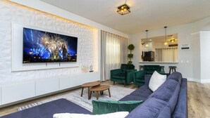 A modern living room with a large flat-screen TV, green upholstered sofas, a small coffee table, and a dining area with a bar. - Windsor Oasis 6BR Pool & Game Room  9 min Disney! (Kissimmee)