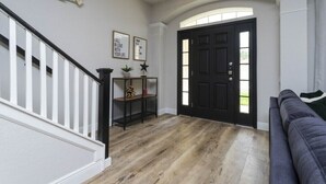 A hallway with a wooden floor, a black door, a staircase with white railings, and a gray sofa. - Windsor Oasis 6BR Pool & Game Room  9 min Disney! (Kissimmee)
