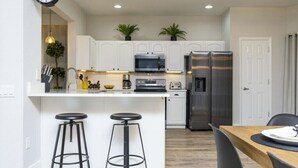 A modern kitchen with white cabinets, a stainless steel refrigerator, a microwave, and two bar stools. - Windsor Oasis 6BR Pool & Game Room  9 min Disney! (Kissimmee)