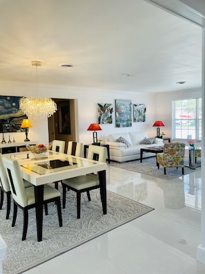 A modern living room with a dining area, featuring a glass-top table, white sofas, and a chandelier.