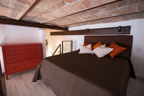 Un dormitori amb sostre de maó, fusta de capçal de llit, una petita còmoda vermella i un llit amb roba de llit marró i almoades taronges.