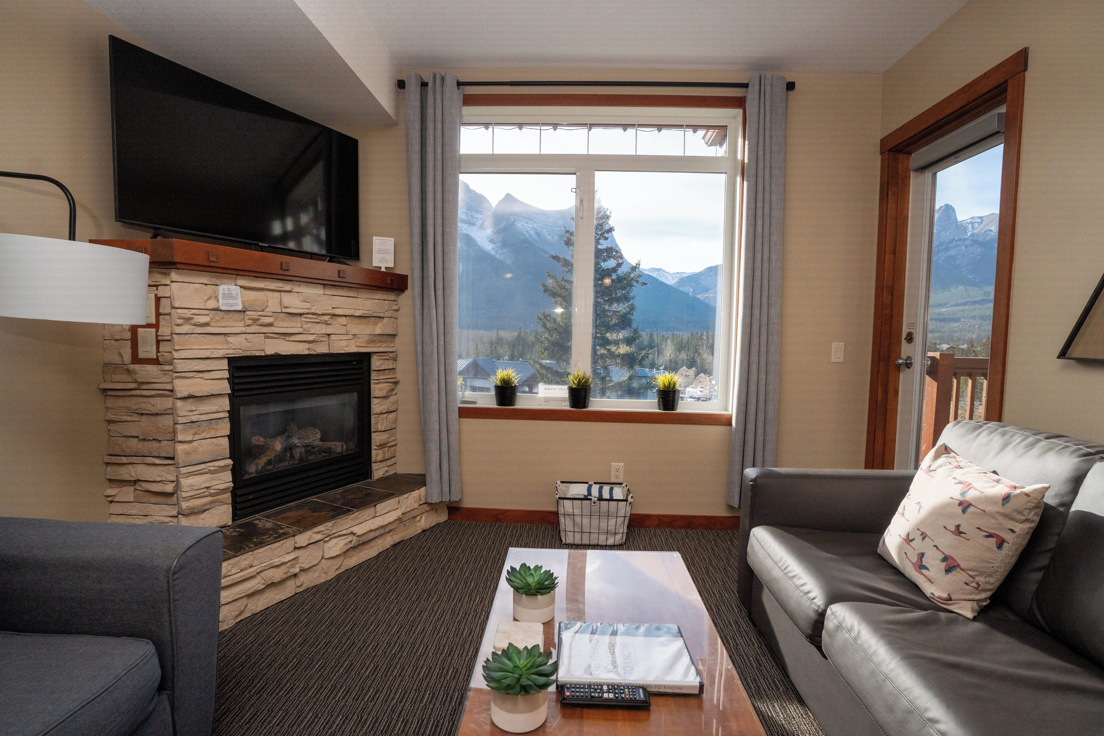 Ein Wohnzimmer mit Kamin aus Stein, einem flachbildfernseher darüber, einer Couch mit Kissen, einem Couchtisch mit Fernbedienung und Buch sowie einem Fenster mit Blick auf die Berge.