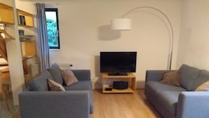 Un saló amb dos sofàs grisos, una televisió de pantalla plana sobre un suport de fusta, una làmpada de peu i una estanteria.