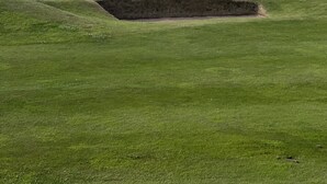 A golf course with a bunker in the foreground, a clear sky, and the ocean in the background.