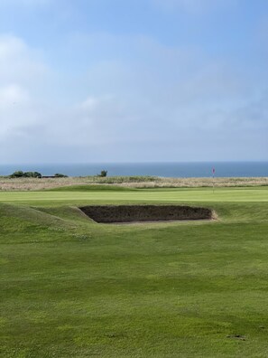 Un terrain de golf avec un bunker au premier plan, un ciel dégagé et l’océan en arrière-plan.