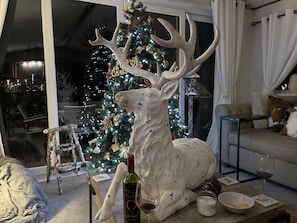 Un salon décoré d’un sapin de Noël, orné d’une grande sculpture de cerf, avec une bouteille de vin et une bougie.