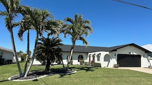 A single-story house with a white exterior, a dark garage door, and a well-maintained lawn with palm trees. - Love Water? Gulf Access, Boat Dock, Private Heated Pool in Lanai. (Cape Coral)