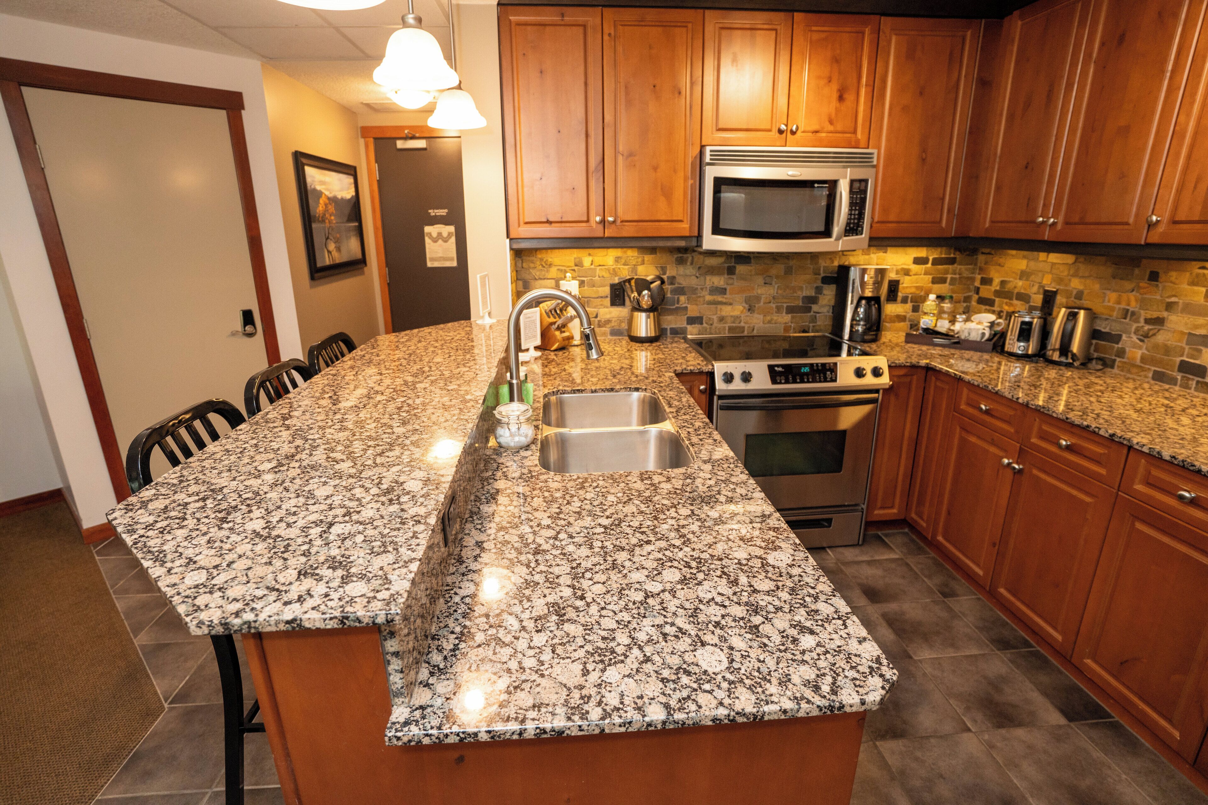 A kitchen with granite countertops, wooden cabinets, a stainless steel sink, a microwave, a stove, and a toaster.