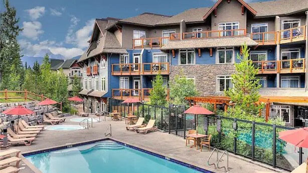 A hotel with a swimming pool, outdoor seating, and a mountain backdrop. - Enjoy a Cozy Condo and Lots of Fun with Family Friendly Pool and Hot Tubs  (Canmore)