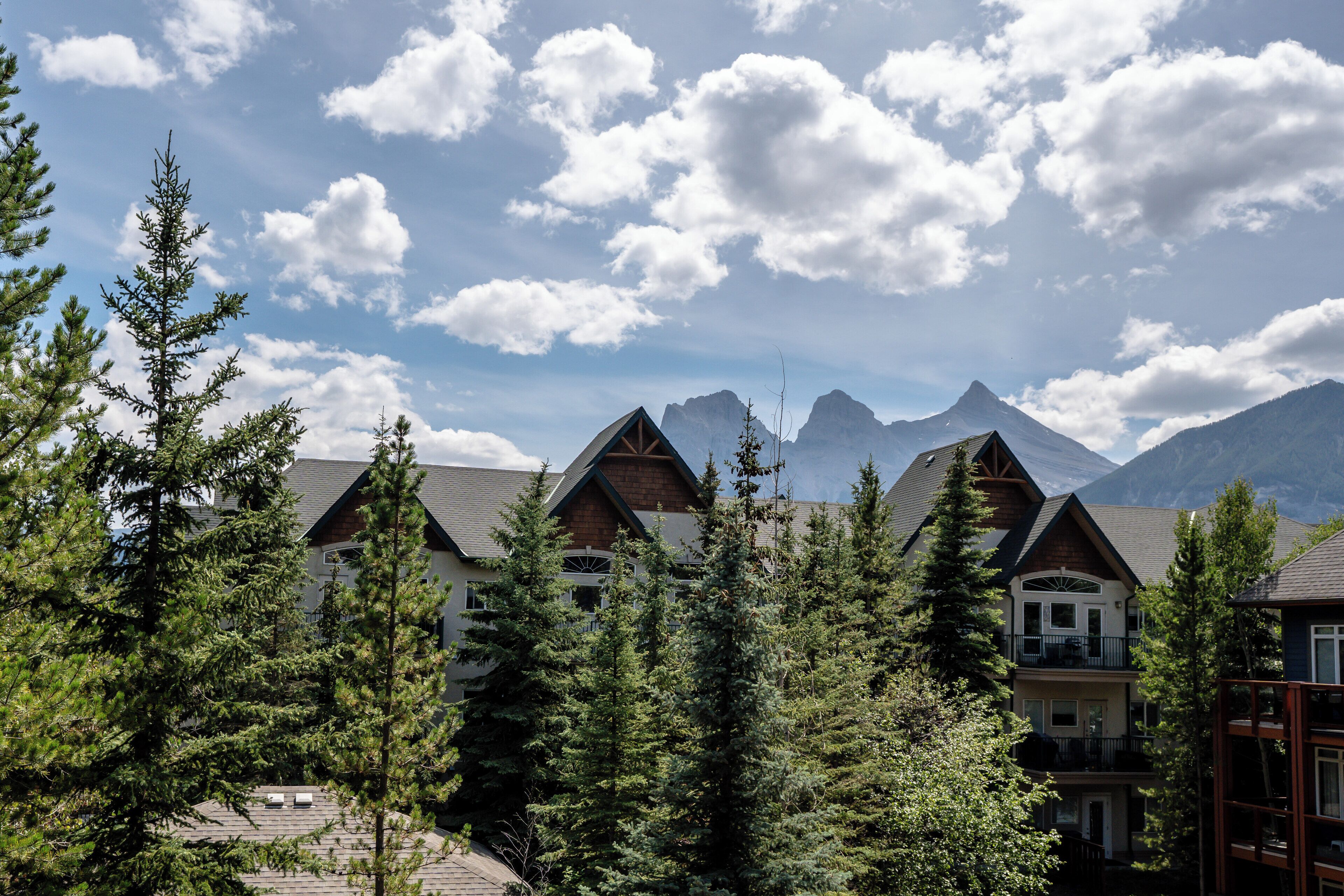 A mountain lodge with multiple buildings, surrounded by evergreen trees and a backdrop of snow-capped peaks.