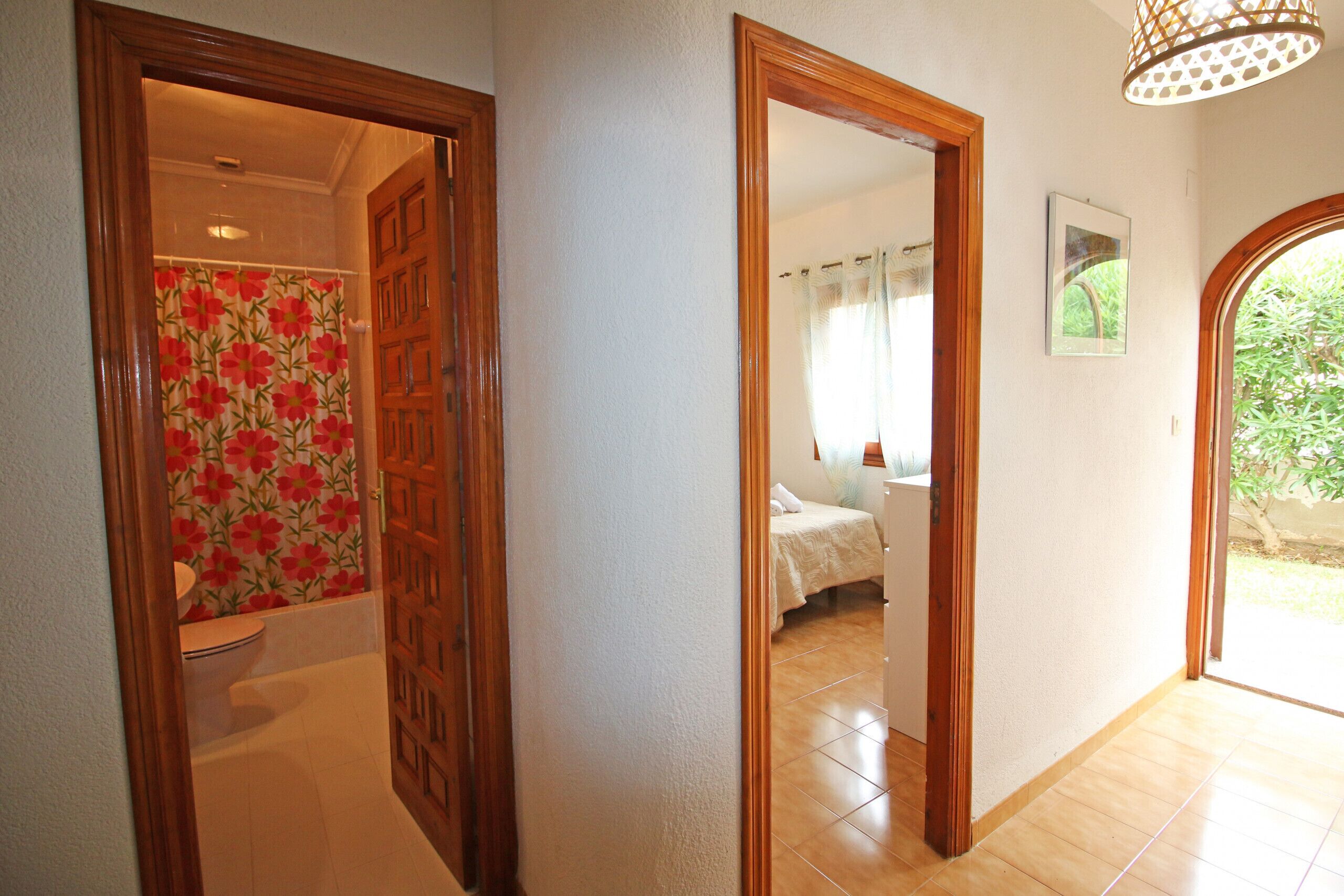 Een gang met een houten deur die leidt naar een badkamer met bloemenbehang, een slaapkamer met een bed en gordijnen, en een grote spiegel die de kamer weerkaatst.