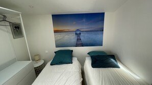 A bedroom with two beds, a wall-mounted picture of a sunset over water, a small bedside table, and a hanging towel rack. - Cozy chef's'apartment in Hyper center... (Andernos-les-Bains)