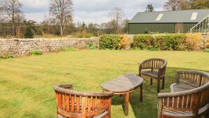 A grassy outdoor area with wooden furniture, a stone wall, and a green building in the background. - Storkery Cottage (Aberdeen)