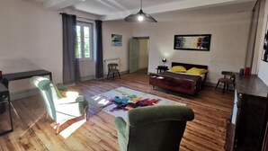 A spacious living room with a wooden floor, a sofa, a chair, a bed with yellow pillows, a radiator, and a ceiling fan. - Maison Gasconne, Charme de L'ancien Allié au Confort du Moderne ! (Saint-Jean-le-Comtal)