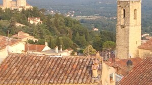 Vista de uma cidade com uma torre imponente, edifícios com telhados de terracota e montanhas ao fundo.