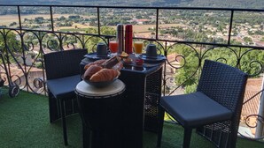 Uma varanda com cadeiras de vime, uma mesa com croissants, suco e uma garrafa térmica. Uma vista panorâmica de uma cidade e colinas é visível pela grade.