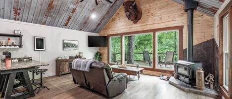 A rustic cabin interior with a stone fireplace, a leather sofa, a dining table, and large windows offering a view of the forest.