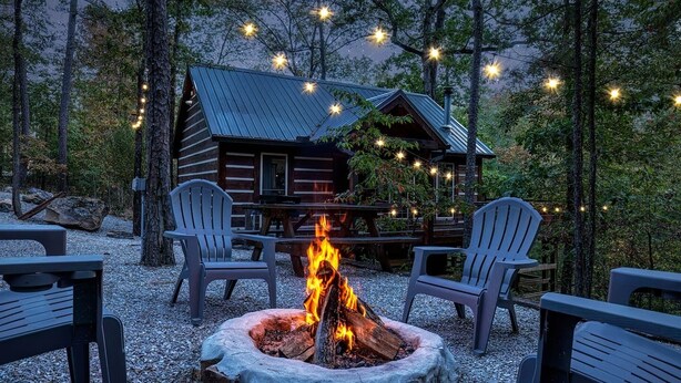 A rustic cabin with a fire pit in the center, surrounded by Adirondack chairs, and string lights overhead. - Runaway Train Cabin at Fox Pass Cabins (Garland County)