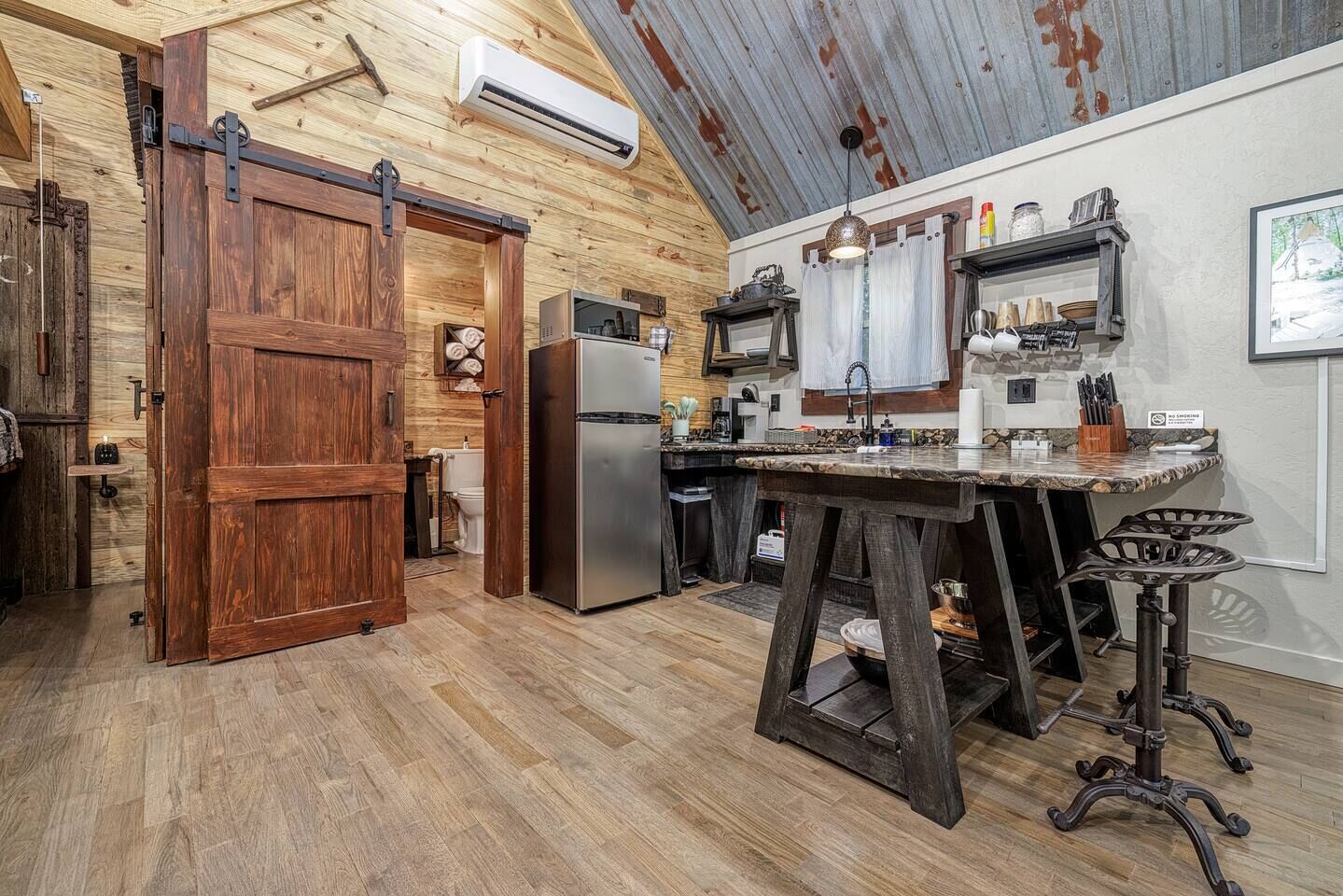 A rustic kitchen with wooden floors, a large wooden door, a refrigerator, a sink, and bar stools.