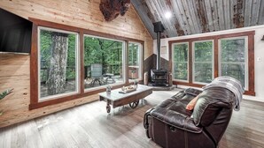 A spacious living room with a large wooden ceiling, a fireplace, a leather sofa, and large windows offering a view of the forest. - Runaway Train Cabin at Fox Pass Cabins (Garland County)