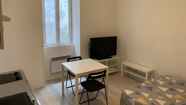A small, clean room with a white table, a black chair, a TV, and a bed with a geometric-patterned bedspread. - Studio Proche Centre Ville (Bayonne)