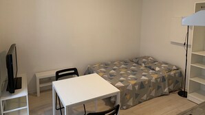 A small, neatly organized room with a bed, a white desk, a chair, and a TV on a stand. - Studio Proche Centre Ville (Bayonne)