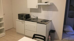 A compact kitchen with a microwave, sink, and stove. There is a chair and a trash bin. - Studio Proche Centre Ville (Bayonne)