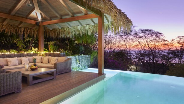 Un área de estar al aire libre techada con piscina, muro de piedra y plantas tropicales.