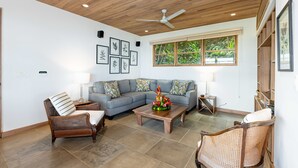 Una sala de estar con techo de madera, un ventilador de techo, un sofá seccional gris, dos sillas de mimbre, una mesa de centro de madera con un centro de mesa floral y una ventana con vista a follaje.