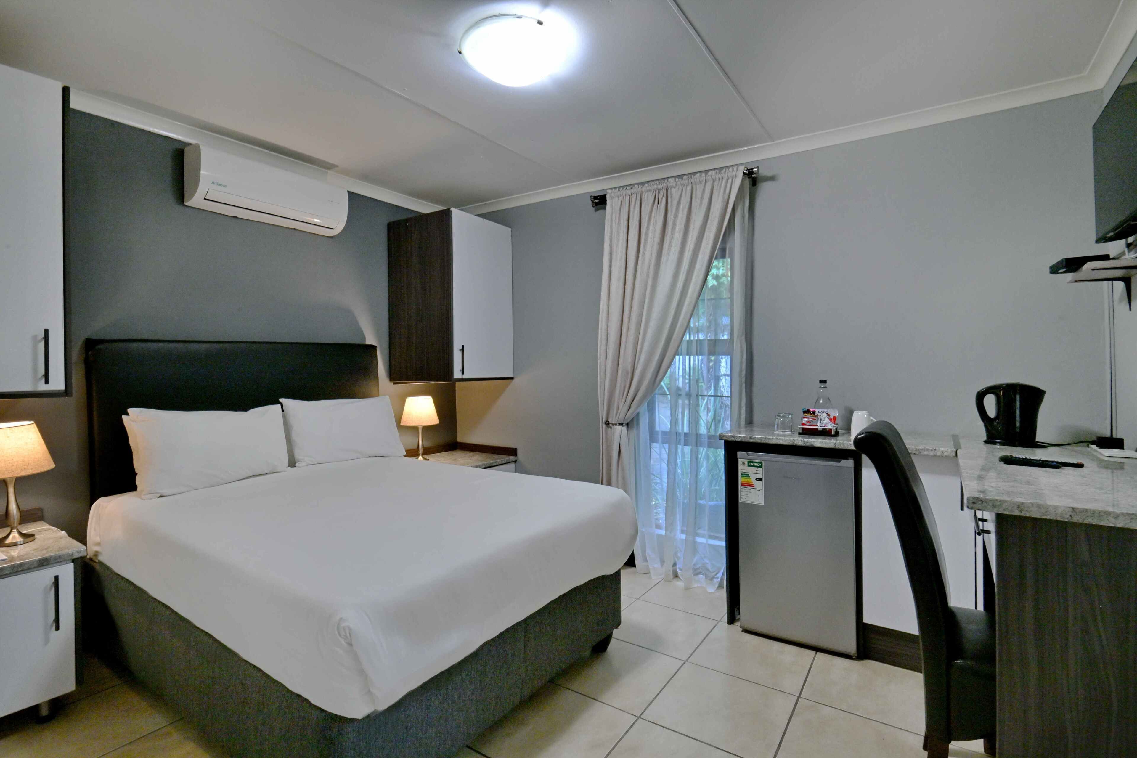 A hotel room with a bed, bedside tables, a desk, a chair, a small refrigerator, and an air conditioning unit.