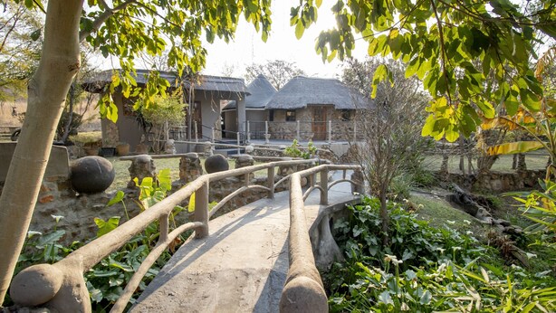 A rustic bridge leading to a stone building with a thatched roof, surrounded by lush greenery and a small stream. - BON Hotel Rustenburg (Rustenburg)