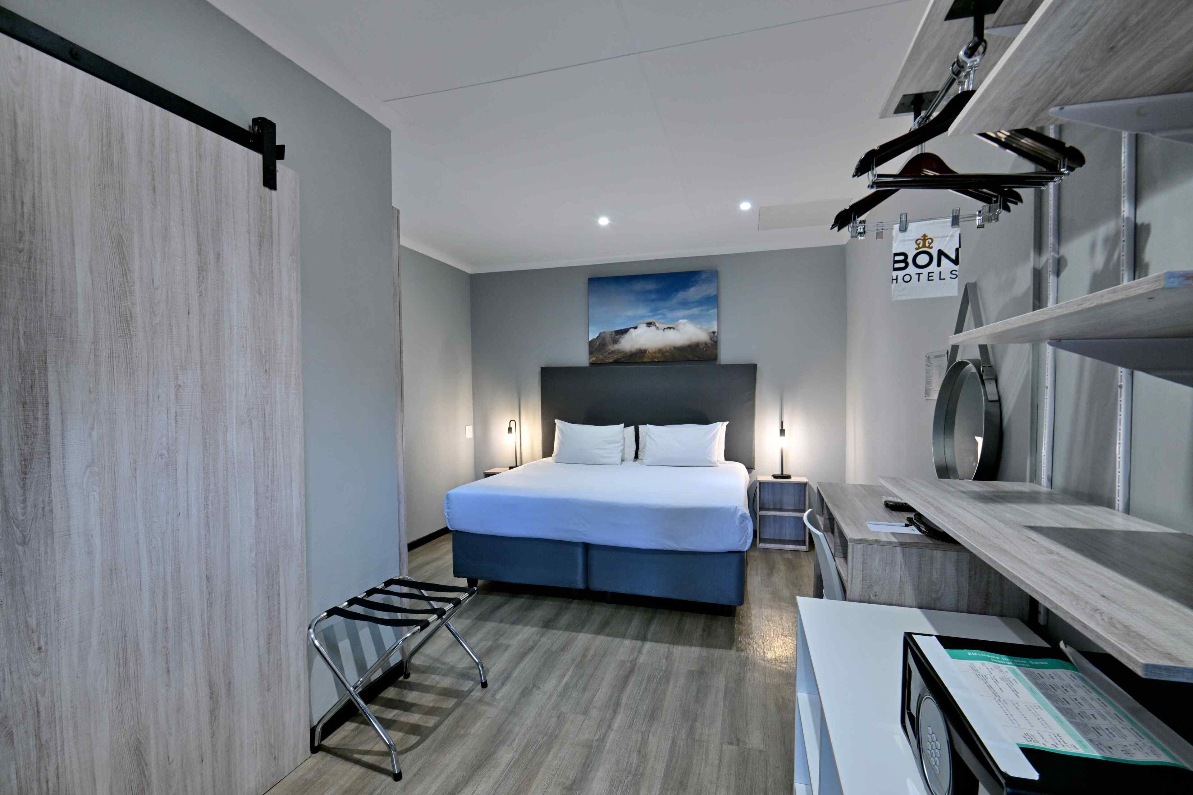 Quarto de hotel moderno com uma cama, uma escrivaninha, uma cadeira, uma mesinha de cabeceira e uma prateleira fixada na parede.