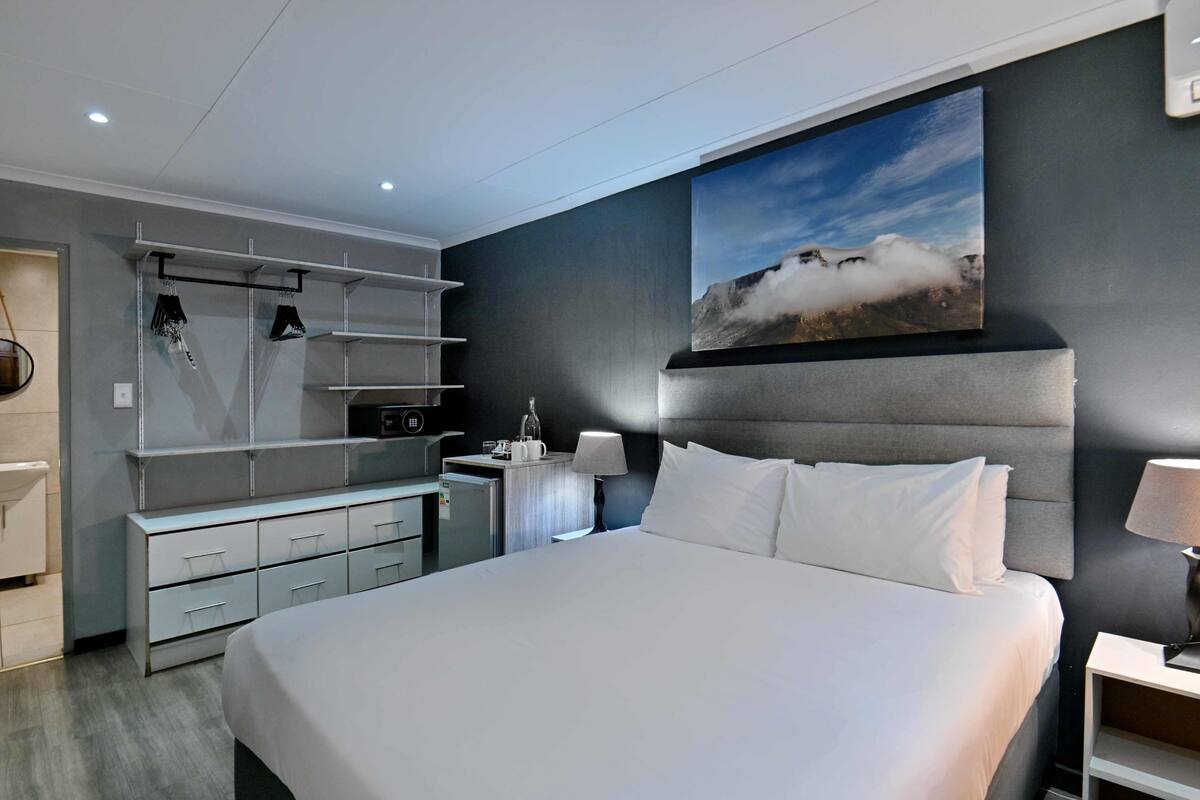 A modern hotel room with a large bed, a wardrobe with hanging space, a desk with a lamp, and a painting of a mountain landscape.